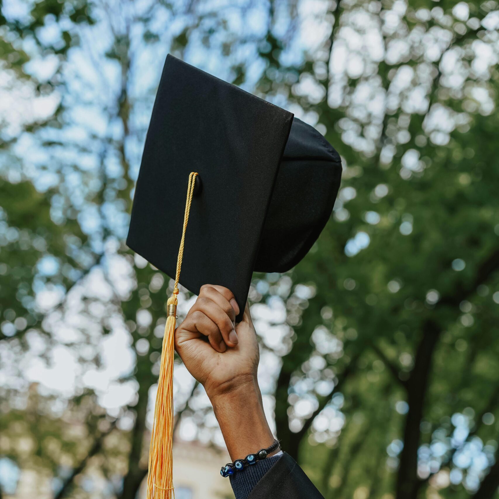 A close-up of a hand raising a graduation cap in celebration among trees.